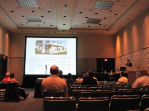 Photo of Mark Taylor speaking to an audience from a podium on a small stage. His presentation is displayed behind him on a large screen.