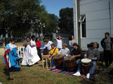 Photo of seven women dancing, with two of them wearing long ruffled skirts. Four men are sitting down, with three of them beating on large drums.