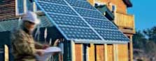 Photo of a man in brown jacket and white hardhat looking at a blueprint. In the background in a large log cabin-style home with photovoltaic panels incorporated on one roof line.