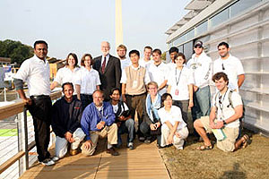 Photo of a group of people next to a white house. Some of the people are standing and others are kneeling, either on a wooden deck or grass. The Washington Monument rises in the background against a clear blue sky.