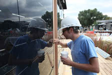 Photo of a man inspecting a small crack between a photovoltaic array and a wooden trim piece. He is wearing a hard hat with "Chris Black" written on it. Construction can be seen in the background.